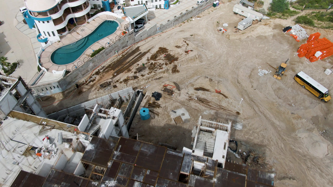 High drone view in San Jose del Cabo, Mexico with the curved pool at upper left, tire-marked dirt across the center, orange materials at right, and no visible people.