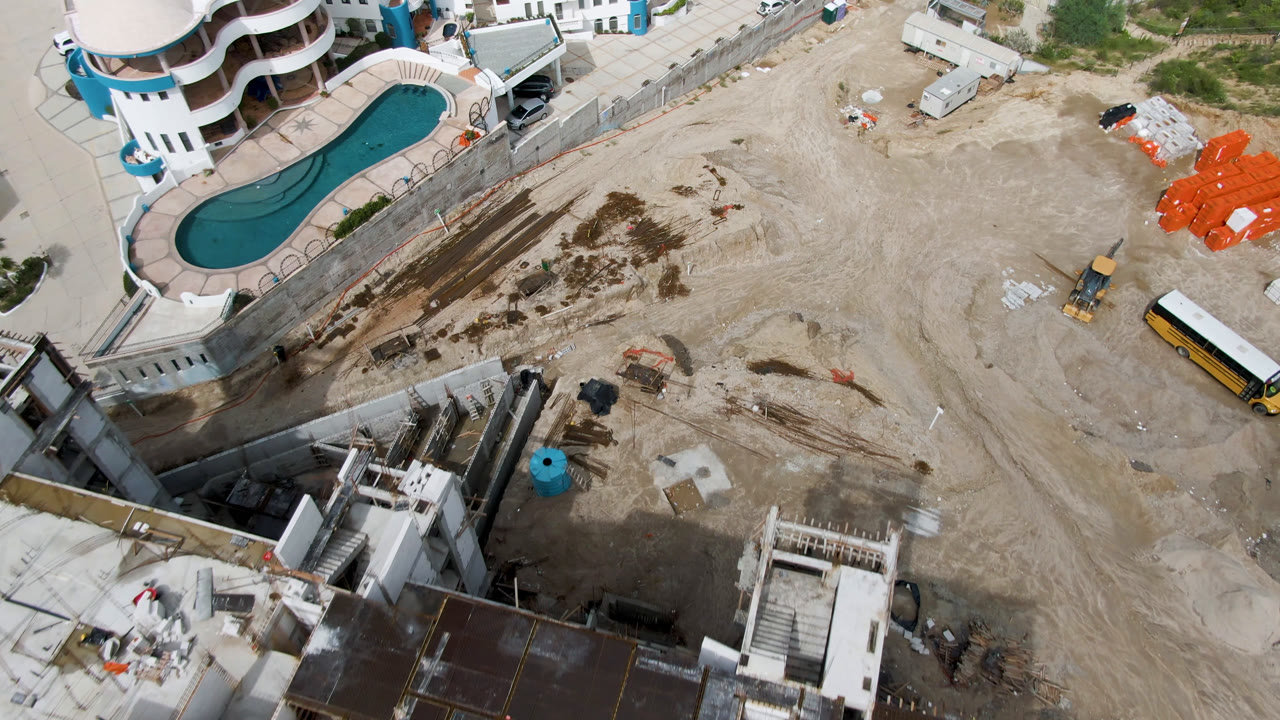 Overhead San Jose del Cabo frame showing the pool and rounded building at left, unfinished concrete work at bottom, a yellow bus near the right edge, and no visible people.