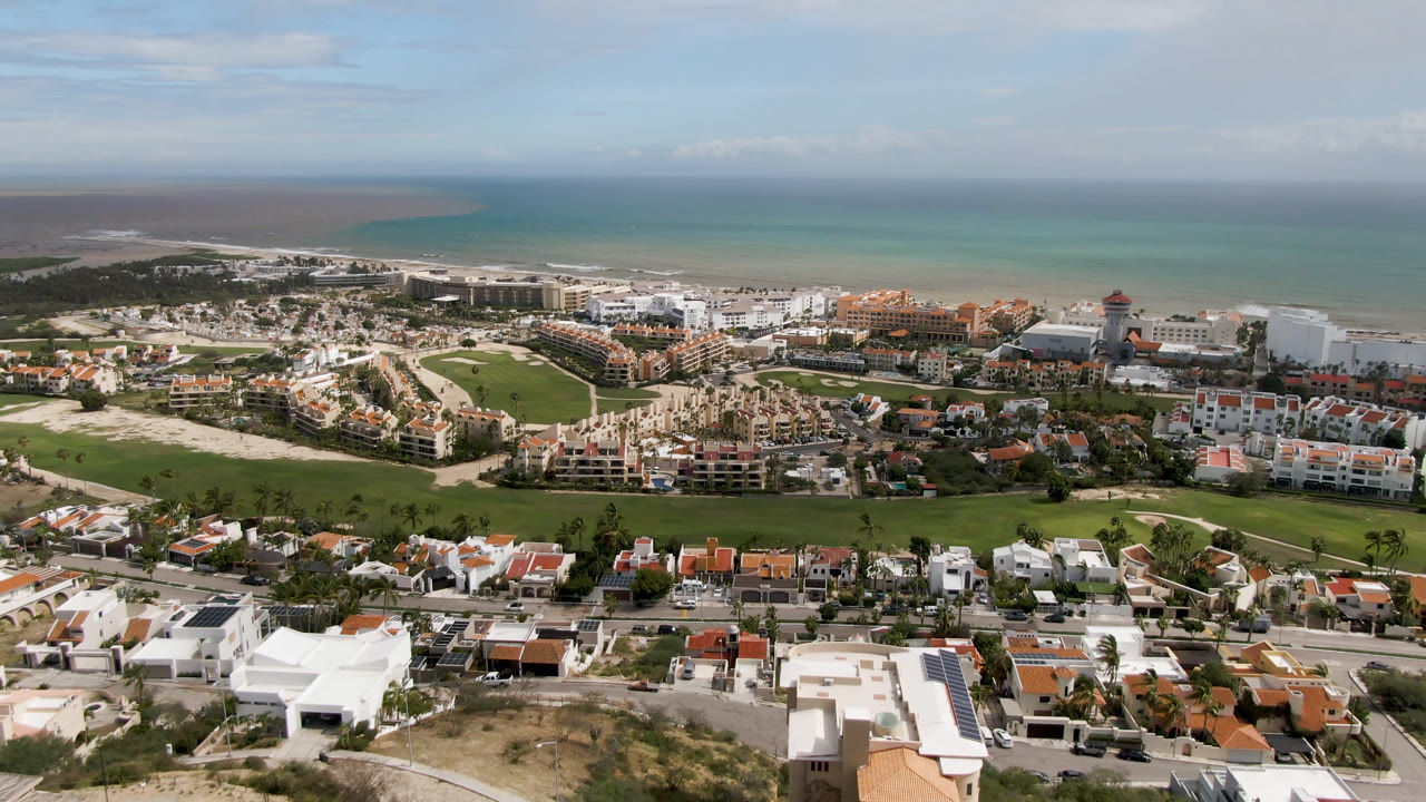 Second aerial frame in San Jose del Cabo showing a broader band of ocean above resort blocks, golf greens, villas, and no visible people.