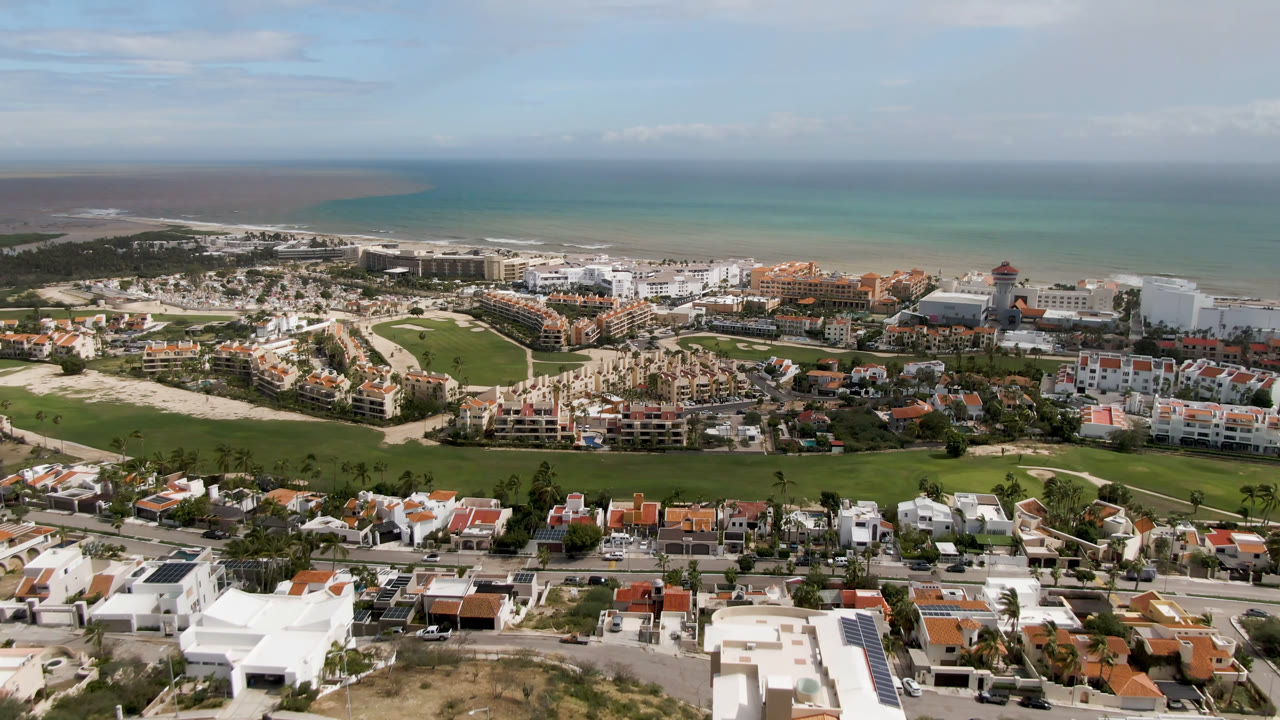 Third San Jose del Cabo aerial frame with the golf course centered below the resort strip, residential roofs in front, ocean behind, and no visible people.