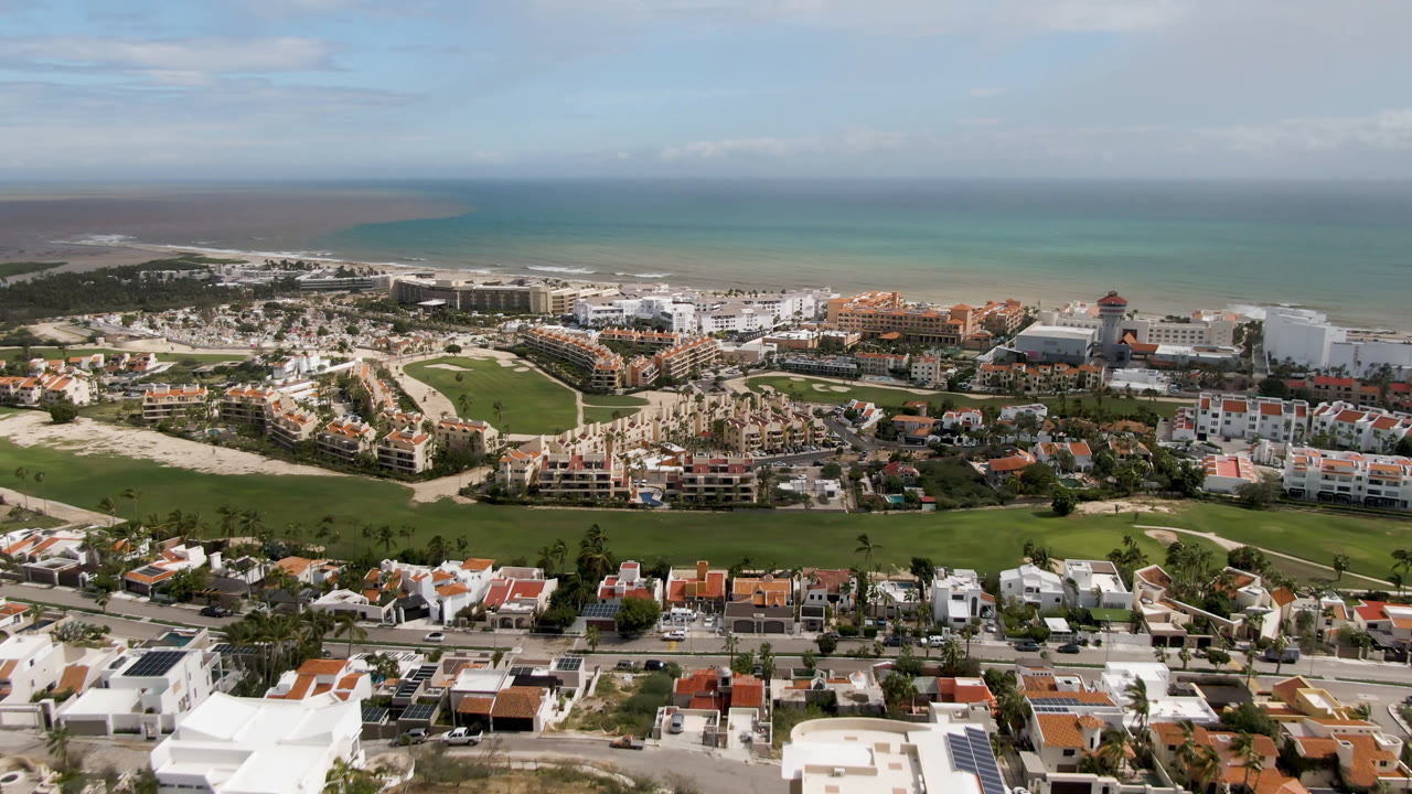 Aerial coastal resort scene with golf course, villas, sandy lots, and ocean horizon under light clouds.