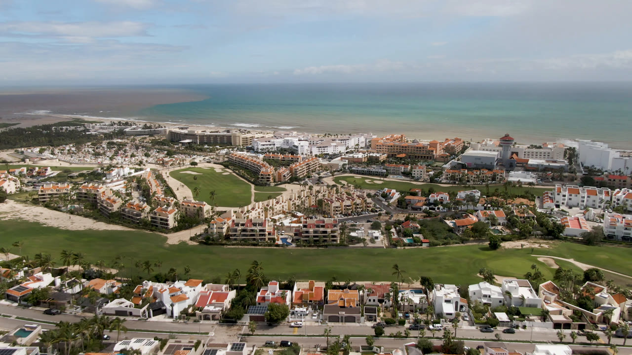 Wide aerial view over San Jose del Cabo showing green golf fairways, resort buildings, beach, turquoise ocean, and no visible people at the opening moment.