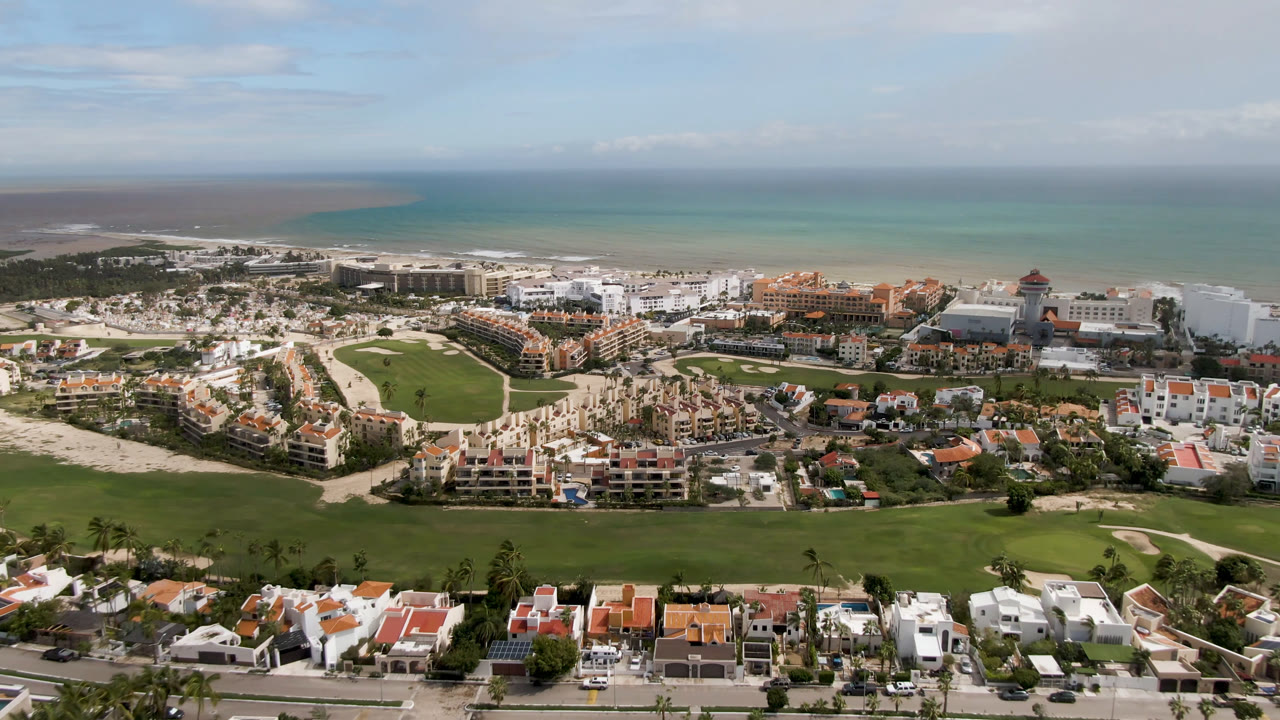 High drone view in San Jose del Cabo with manicured fairways below, clustered homes and resort structures across the middle, beach and ocean in the background, and no visible people.