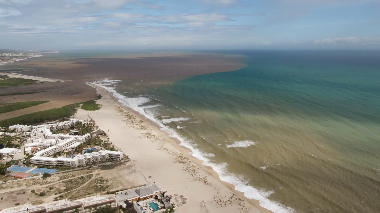 Aerial frame over San Jose del Cabo showing white resort buildings beside a pale sand beach, bands of turquoise water, distant coastline, clouds, and no visible people.