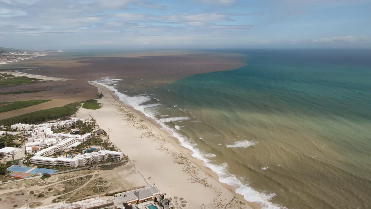 High aerial view in San Jose del Cabo with resort buildings lower left, a diagonal strip of sand, foamy surf lines, green-blue ocean water, and no visible people.