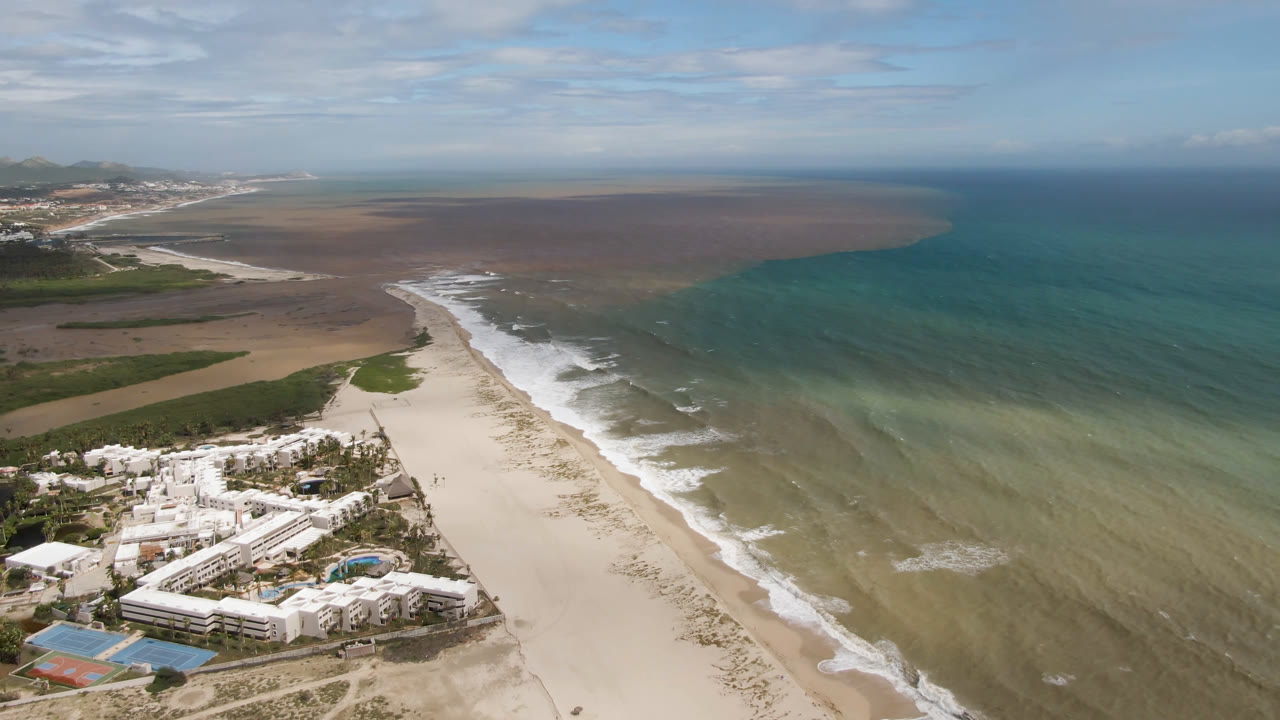High aerial view in San Jose del Cabo, Baja California Sur, showing a white beachfront resort below, a long sandy beach, green-blue water, distant hills, and no visible people.