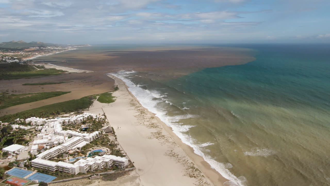 Wide aerial coastline with resort buildings, beach, surf, and open water.