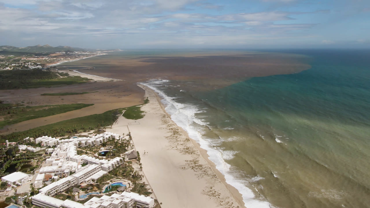 Aerial frame over San Jose del Cabo showing white resort buildings in the lower left, a wide sandy beach, surf bands, turquoise ocean, distant hills, and no visible people.