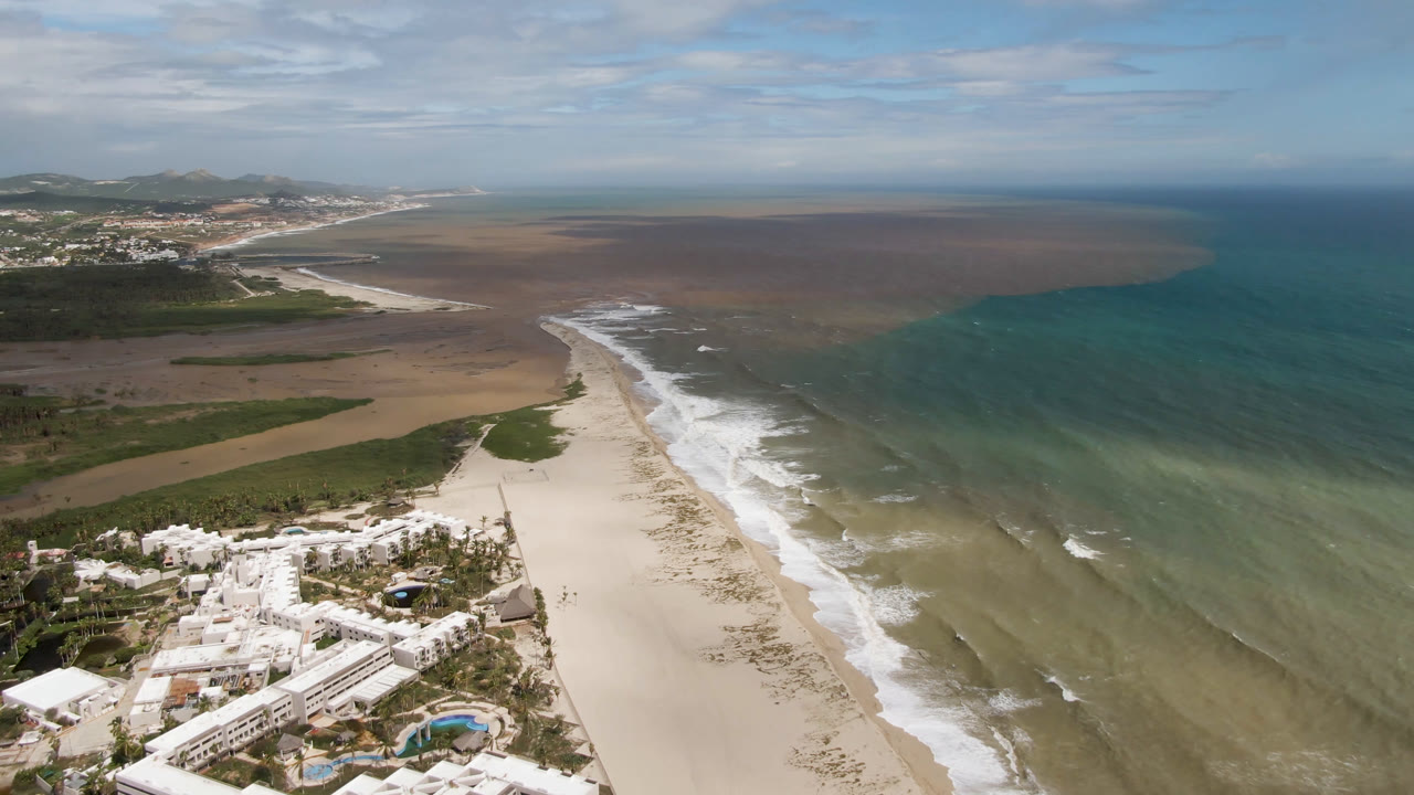 Aerial view in San Jose del Cabo with resort rooftops at the bottom, pale sand through the middle, breaking white waves, mixed turquoise and brown water, and no visible people.