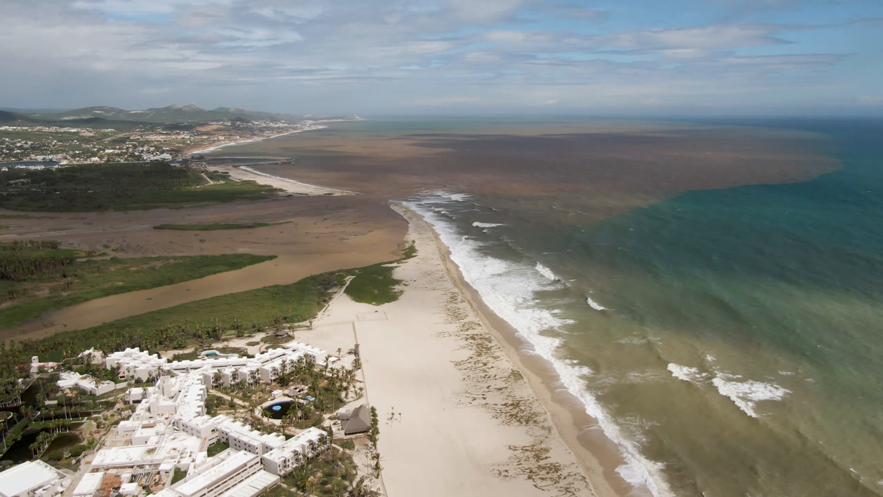 Aerial frame from San Jose del Cabo, Mexico showing white buildings at the lower left, a broad sandy beach, surf lines, colored ocean water, distant town, hills, clouds, and no visible people.