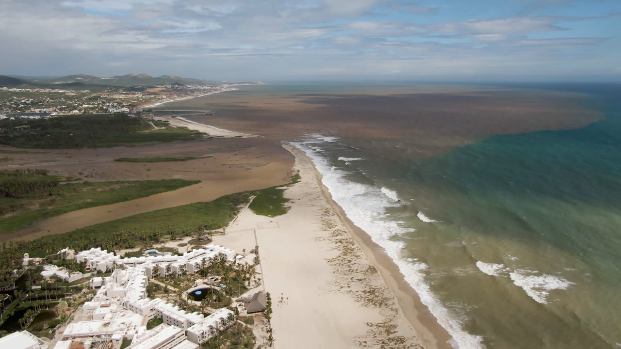 Aerial view in San Jose del Cabo, Mexico with white buildings closer to the bottom edge, a long pale beach, broken wave lines, blue-green ocean, inland fields, distant settlement, and no visible people.