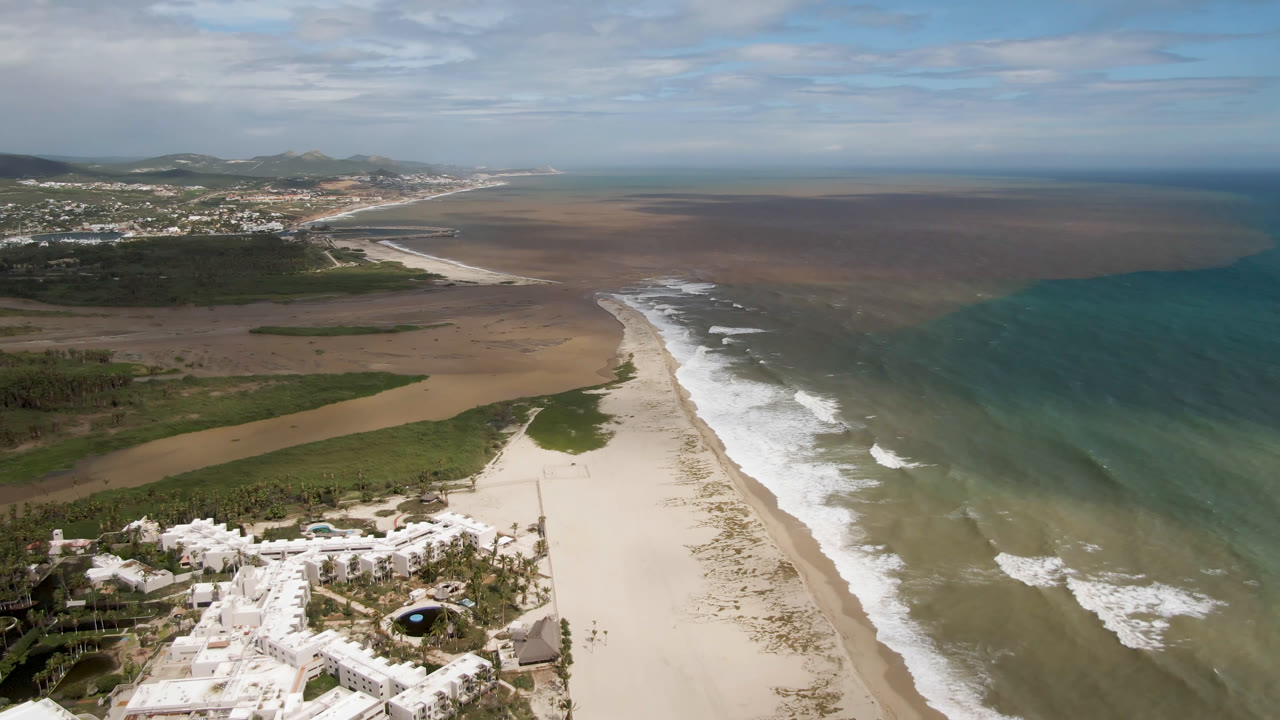 Wide coastal aerial with beach, surf, ocean, and town in the distance.