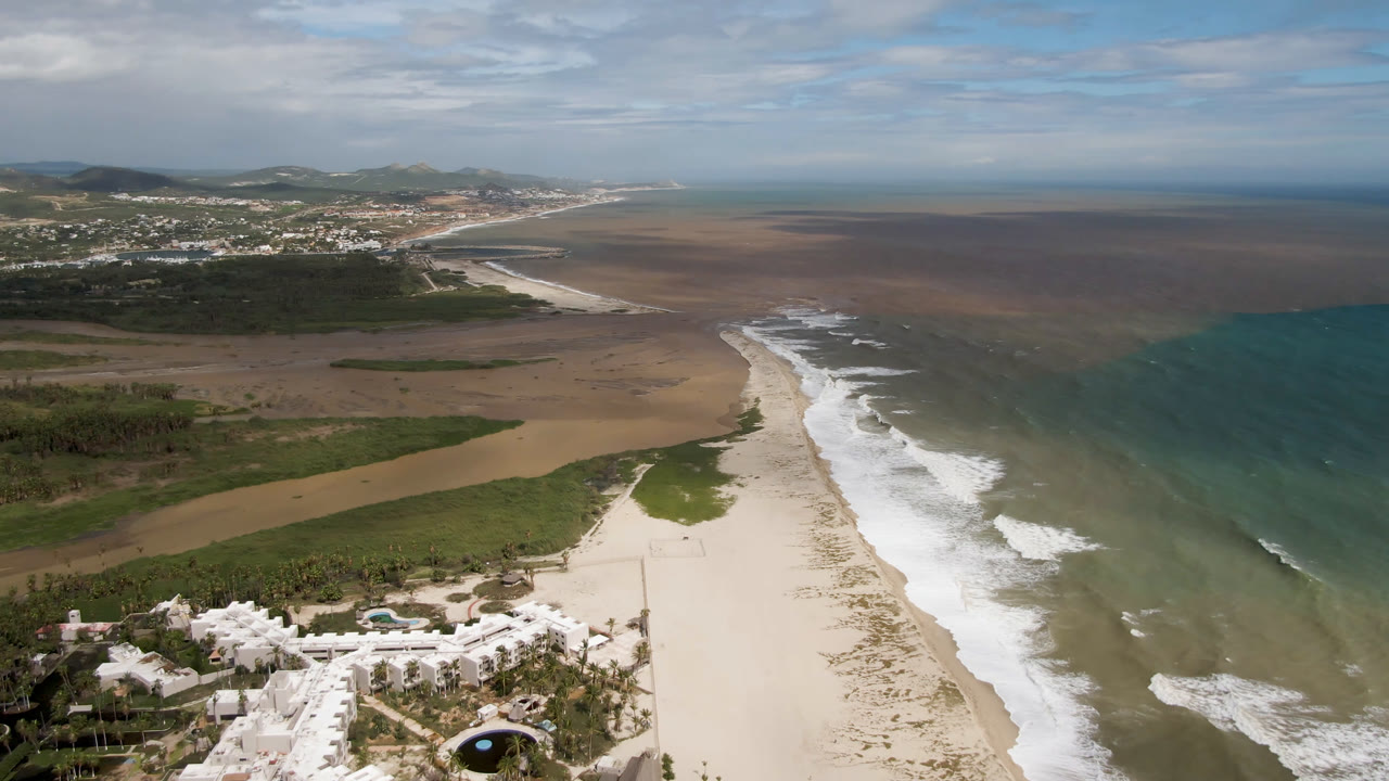 High aerial frame over San Jose del Cabo showing a sandy beach below, white surf along the shore, mixed turquoise and brown ocean water, coastal vegetation, distant hills, and no visible people.