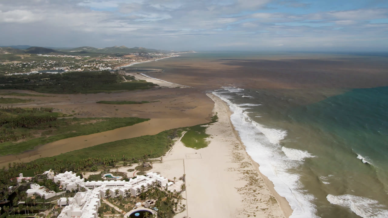 Aerial view in San Jose del Cabo with resort-style buildings at the lower edge, a pale beach running into the distance, surf breaking near center frame, brown water offshore, and no visible people.