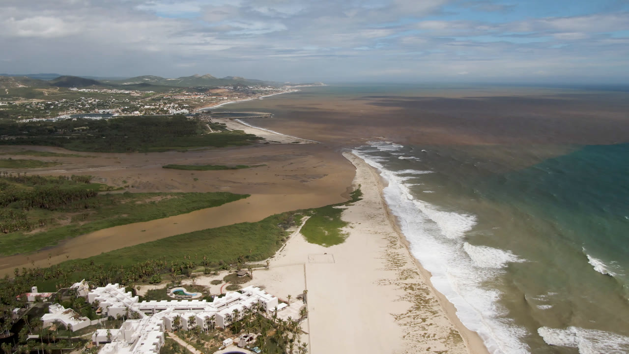 Wide aerial coastline with white surf, sandy beach, green vegetation, ocean, and distant town.