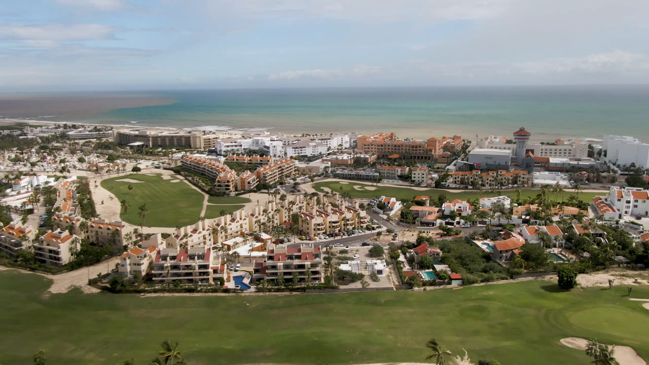 Second frame over San Jose del Cabo shows a broad green fairway, clustered resort buildings, pale roads, ocean water, and no visible people under bright daylight.
