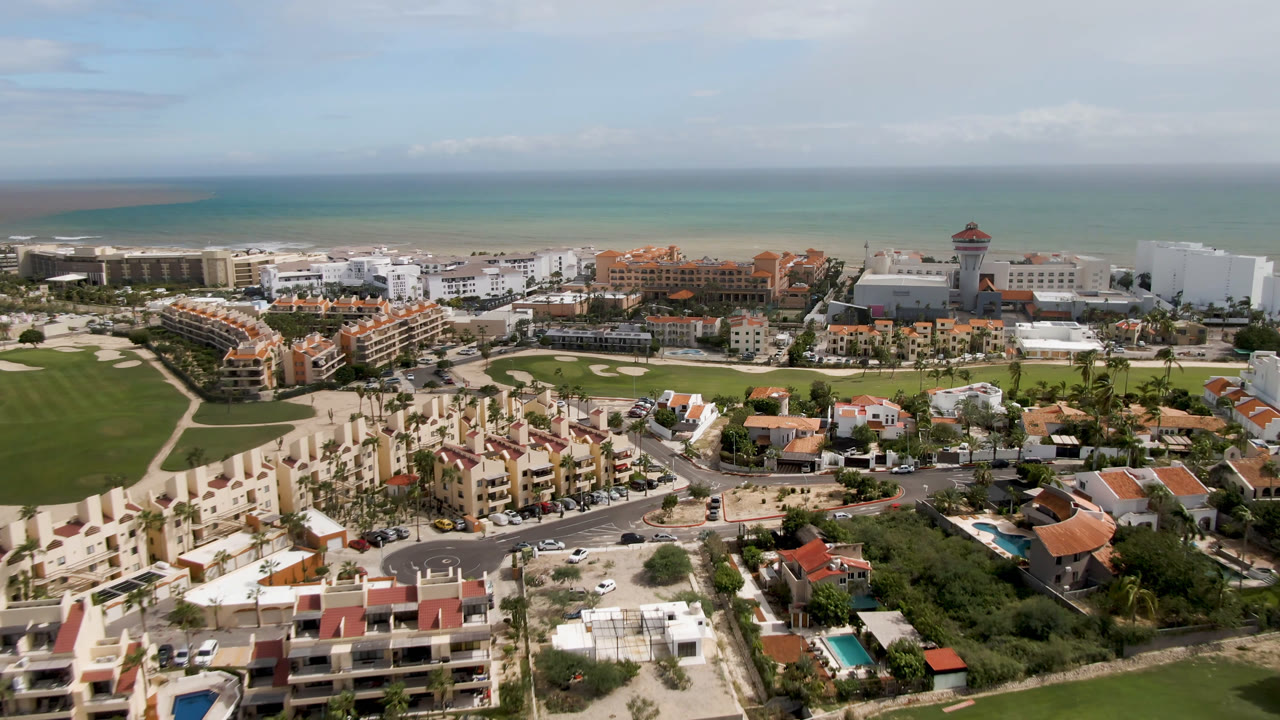 Aerial view in San Jose del Cabo with apartment-style resort blocks, manicured golf greens, foreground homes, a coastal road layout, turquoise water, and no visible people.