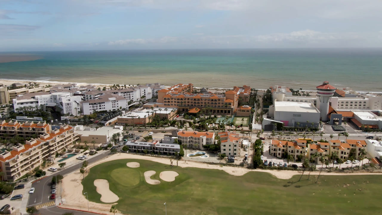 Aerial view over San Jose del Cabo with a golf green and sand traps in front, orange-roofed hotel buildings near the beach, green-blue ocean beyond, and no visible people.