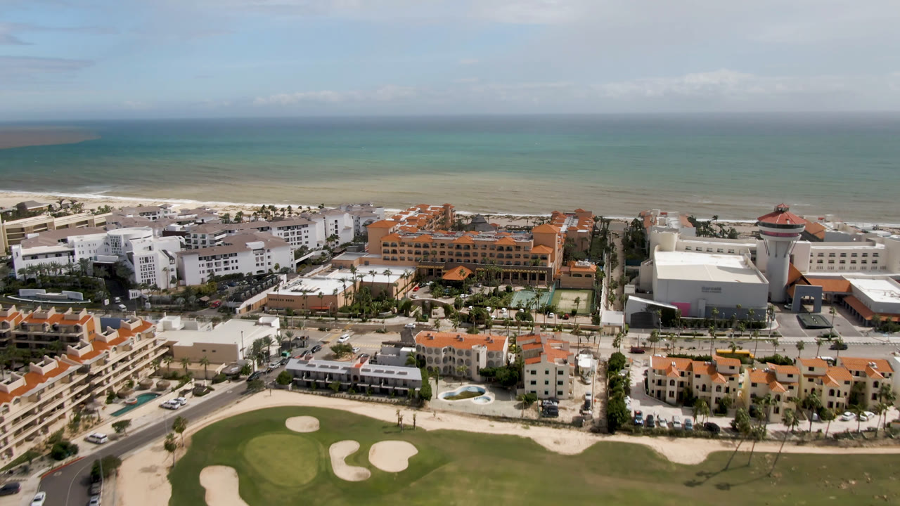 High aerial frame in San Jose del Cabo showing white and orange-roofed hotel blocks, a beachfront road, pale sand, layered shallow water, and no visible people.