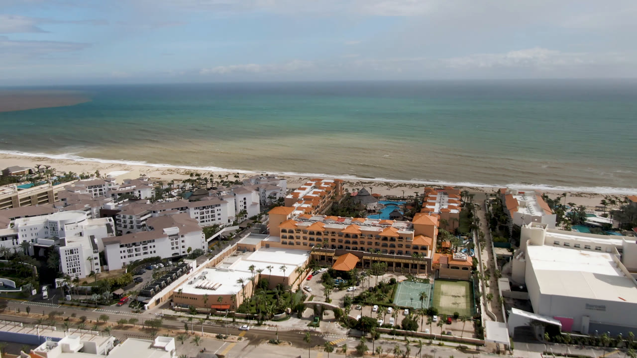 High aerial view over San Jose del Cabo resort buildings, pools, beach, surf, and ocean horizon, with no visible people in the opening frame.