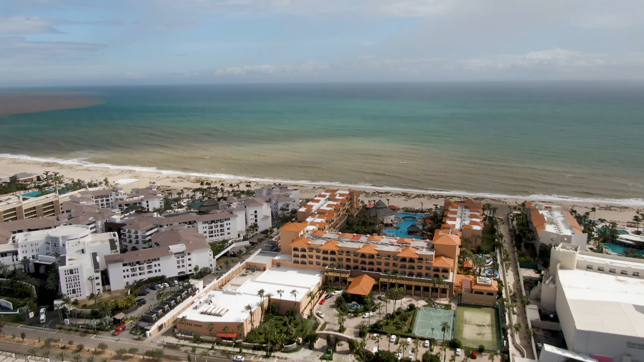 Aerial frame over San Jose del Cabo showing the shoreline, pale surf bands, hotel rooftops, blue pools, and streets, with no visible people.