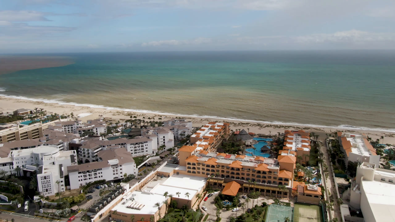 Wide aerial over San Jose del Cabo beachfront hotels, orange-roofed resort sections, pools, beach, and green-blue ocean, with no visible people.