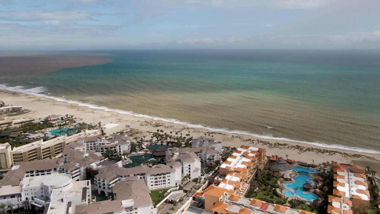 Aerial frame over San Jose del Cabo resort buildings and blue pools beside a pale beach and turquoise ocean, with no visible people.