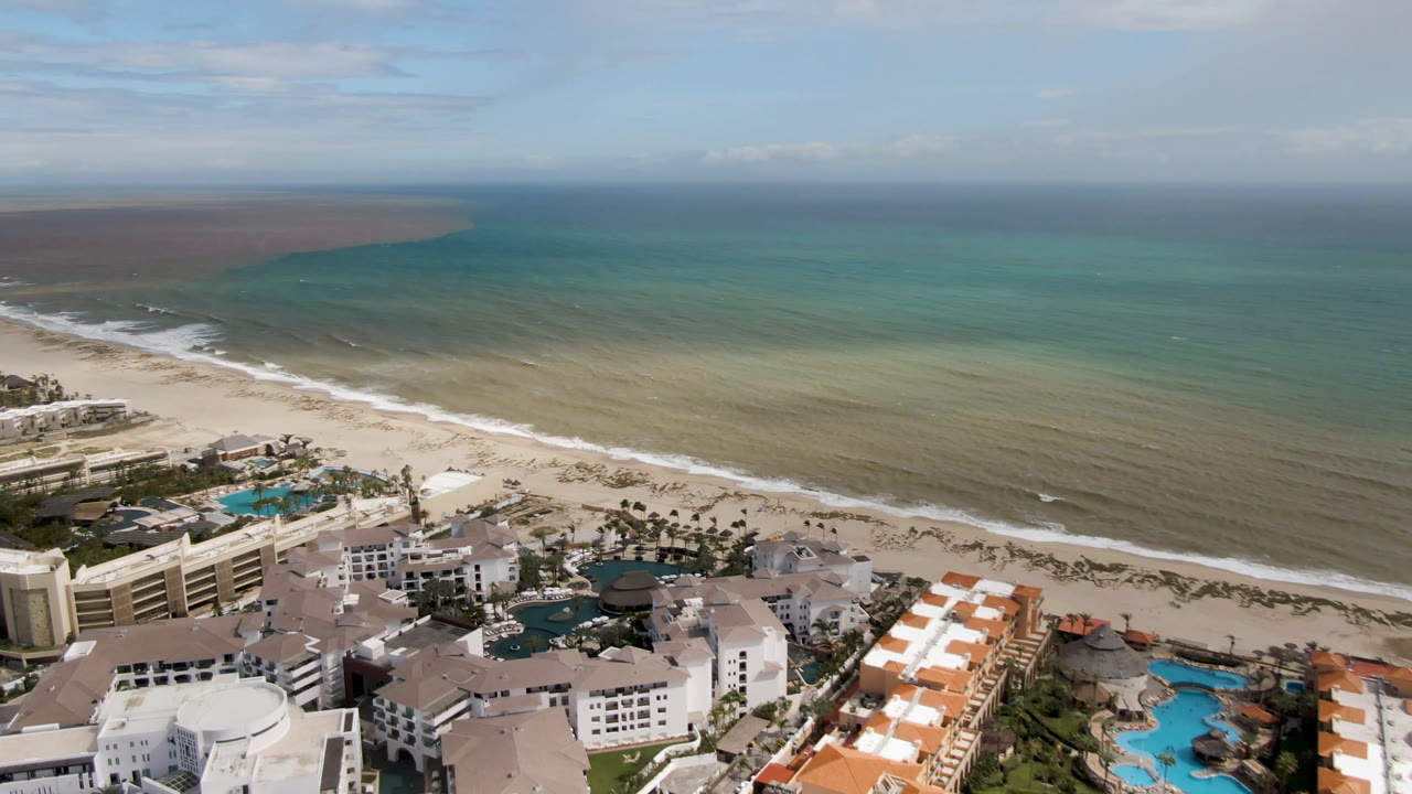 Aerial view in San Jose del Cabo with resort rooftops, turquoise pools, surf along the beach, and a broad ocean horizon, with no visible people.