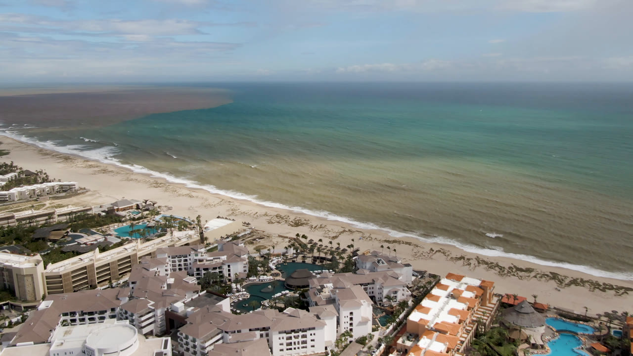 High aerial angle on resort buildings, beach, surf, and open water.