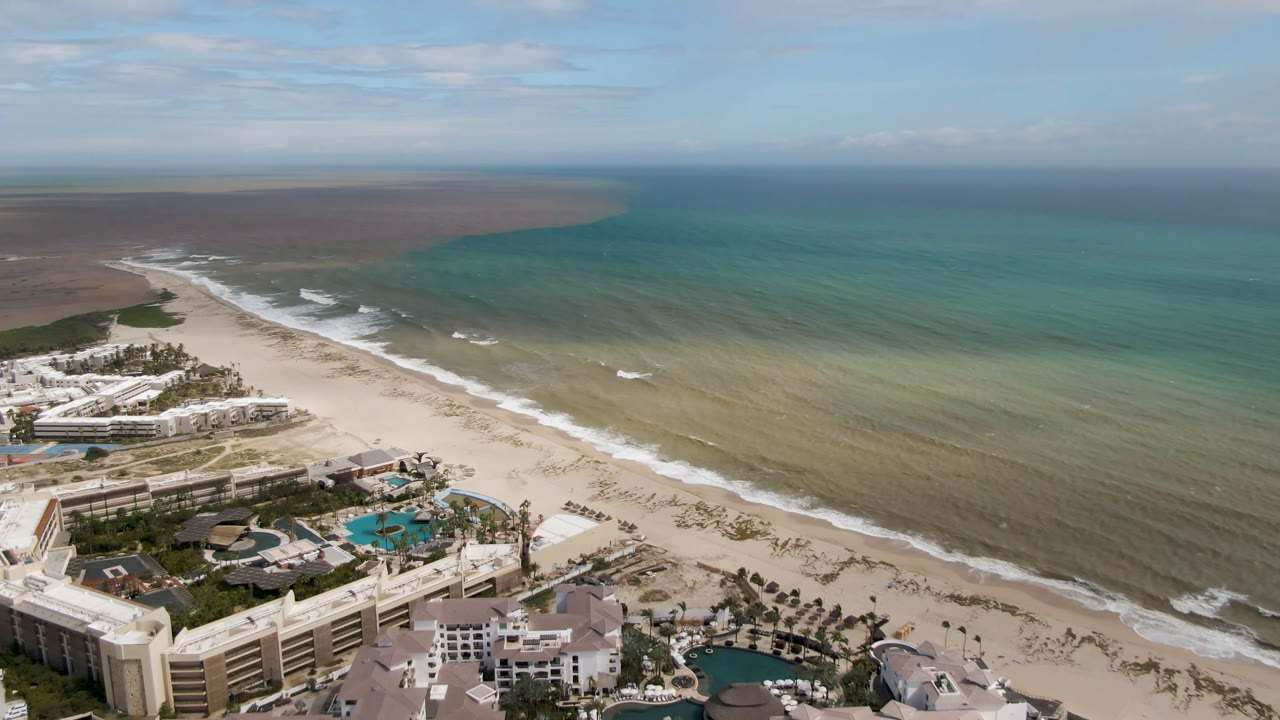 Aerial view over San Jose del Cabo with resort buildings and pools at the lower left, a wide beach, turquoise water, and no visible people.