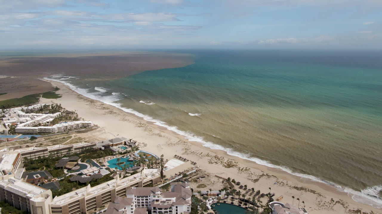 Drone view in San Jose del Cabo looking diagonally along the beach, with white resort blocks, blue pools, rolling surf, and no visible people.