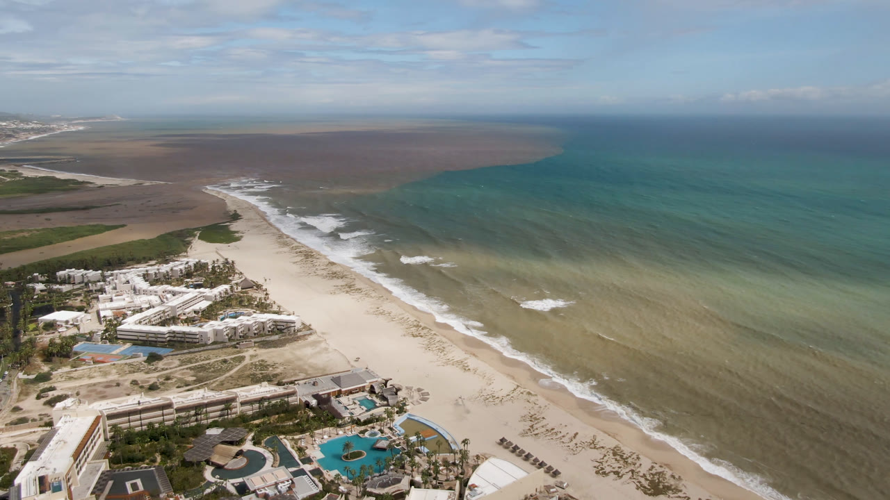 Aerial frame over San Jose del Cabo, Baja California Sur, showing resort pools and buildings at the bottom, a pale beach, turquoise water, brown shallows, and no visible people.