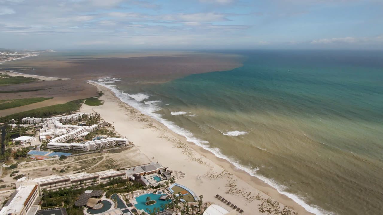 Drone-height view in San Jose del Cabo with white resort buildings and blue pools below, surf tracing the beach, wide green-blue ocean, and no visible people.