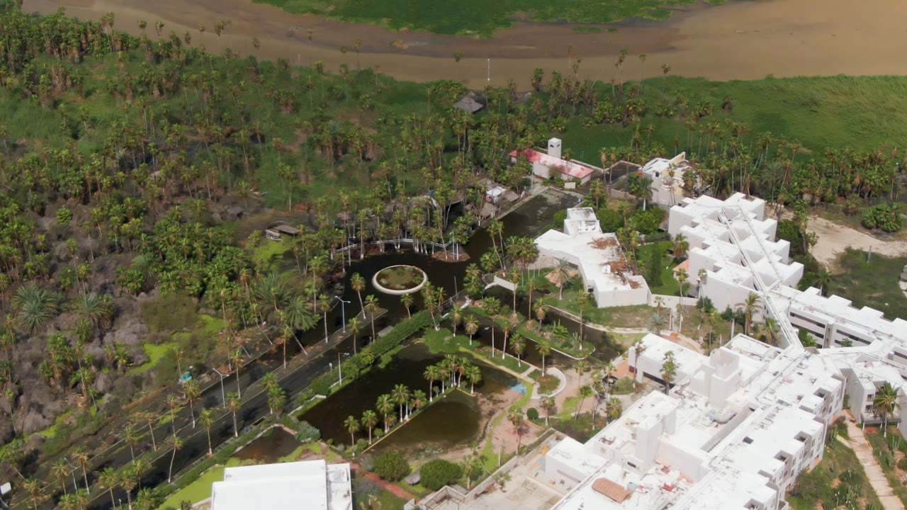 Opening overhead frame in San Jose del Cabo shows white buildings beside dark ponds, curved paths, palm rows, dense green trees, and no visible people.