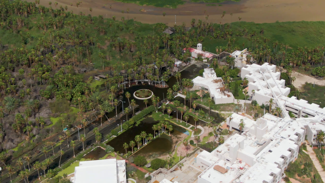 Drone view of a park pond, bridge area, trees, and white architecture.