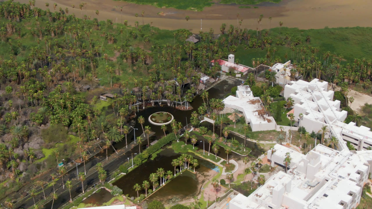Opening aerial frame over a landscaped park in San Jose del Cabo, Mexico, with white angular buildings, dark ponds, palms, curved paths, distant water, and no visible people.