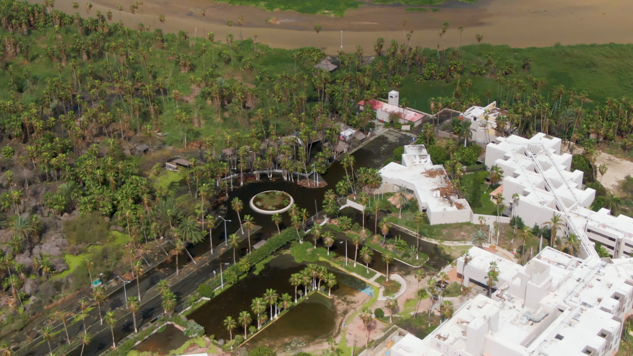 One second into the San Jose del Cabo aerial, the circular fountain and surrounding ponds sit left of white buildings, with palms, paths, green vegetation, and no visible people.