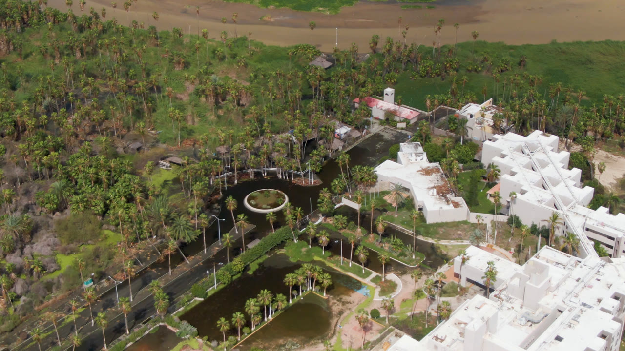 Drone view of landscaped ponds, palms, white roofs, and vegetation near water.