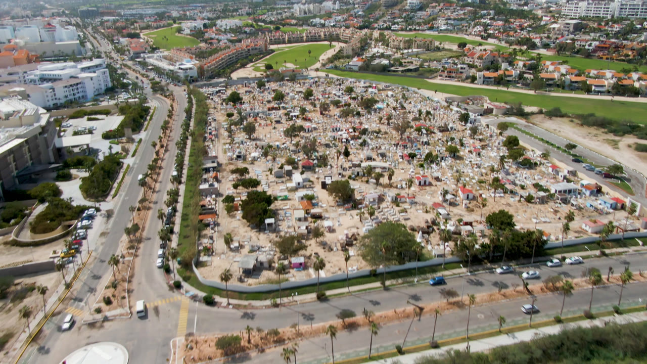 Aerial view of a cemetery beside roads, buildings, a golf course, and distant hills.
