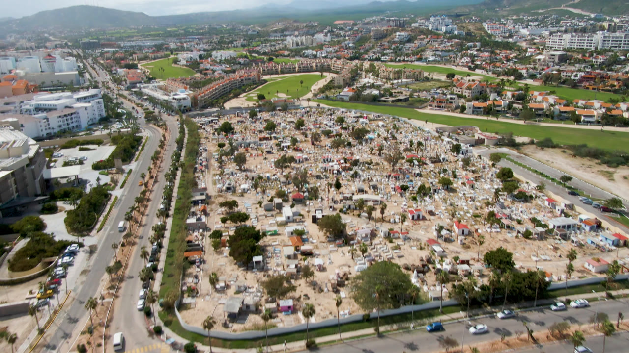 Aerial frame in San Jose del Cabo showing the cemetery filling the lower center, a road corridor with small cars at left, white buildings nearby, golf fairways behind, and no visible people.