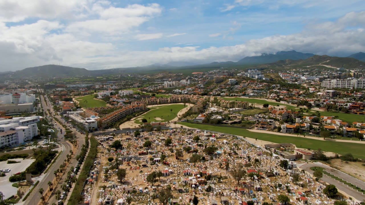 Opening aerial frame over San Jose del Cabo, Mexico, with a crowded cemetery in the foreground, a green golf course beyond it, roads on both sides, mountains in the distance, and no visible people.