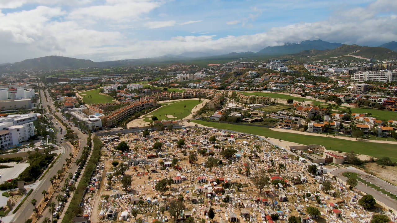Second aerial frame in San Jose del Cabo with cemetery plots across the lower view, curved golf fairways and sand areas behind them, city buildings, distant hills, and no visible people.