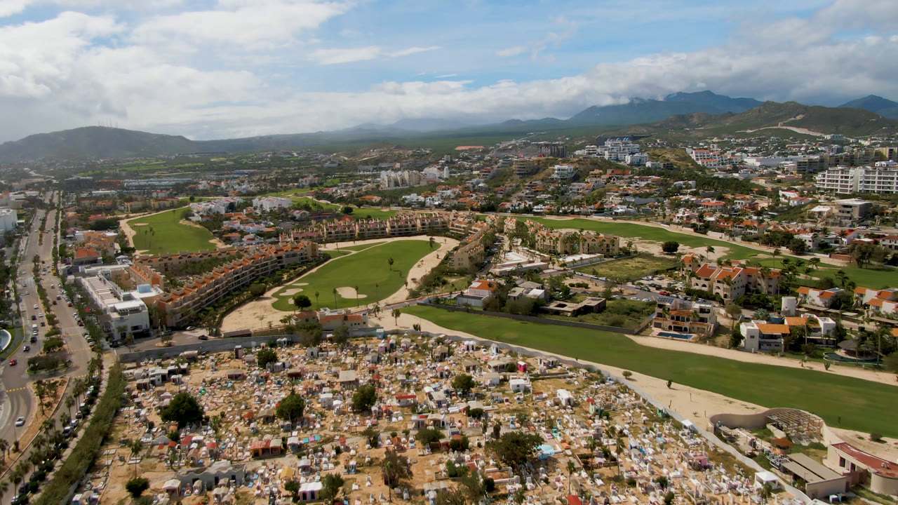 Second aerial frame in San Jose del Cabo showing the cemetery lower in frame, a green golf course with pale paths, residential blocks, a straight road at left, distant mountains, and no visible people.
