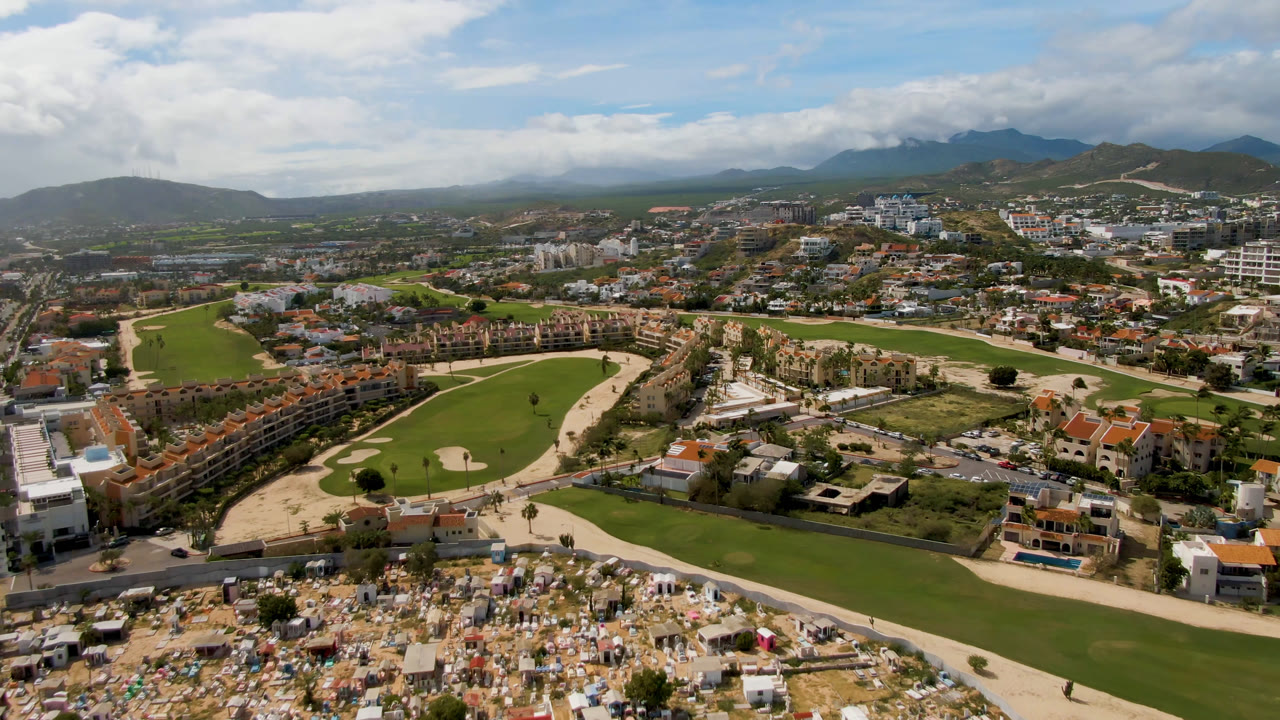 Wide aerial view of a golf course, roads, buildings, hills, and clouds.