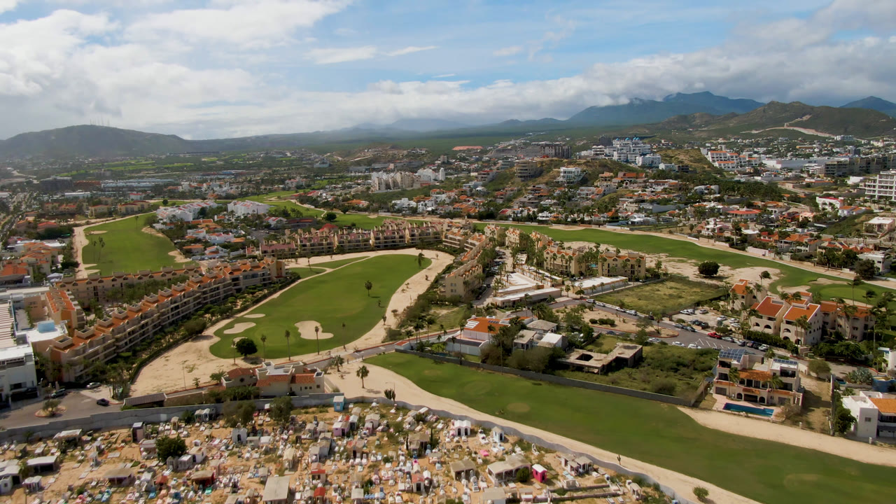 Second aerial frame in San Jose del Cabo showing the fairways and sand traps centered below residential buildings, roads, distant hills, low clouds, and no visible people.