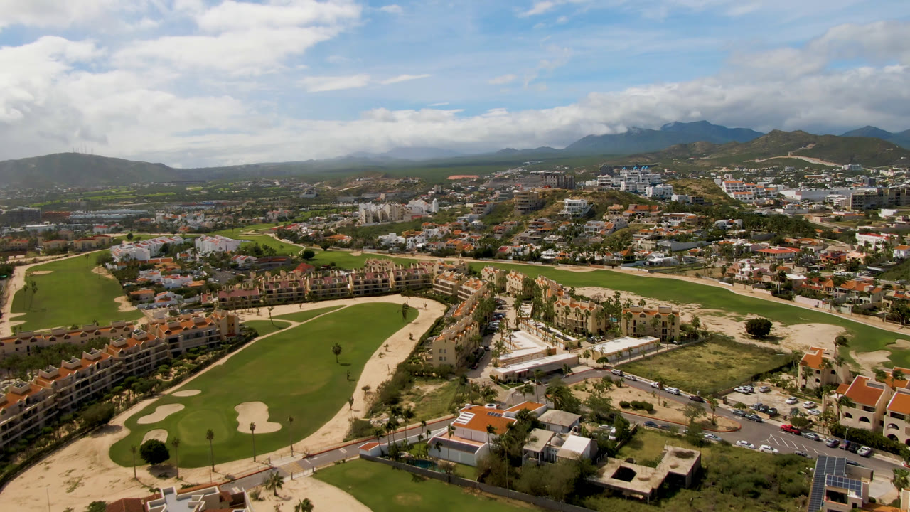 High aerial frame over a San Jose del Cabo golf course with green fairways, sand bunkers, red-roofed buildings, roads, distant mountains, broken cloud, and no visible people.