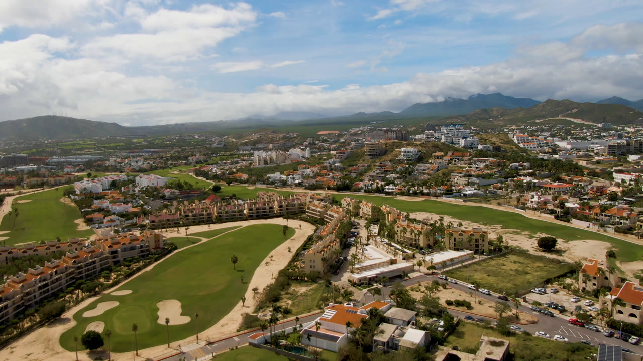Aerial view in San Jose del Cabo showing fairways and pale bunkers in the foreground, clustered buildings and roads in the midground, mountains behind, and no visible people.