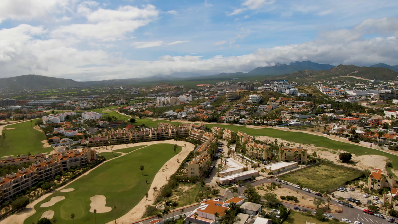 Wide aerial over a green golf course with housing, roads, clouds, and mountains.