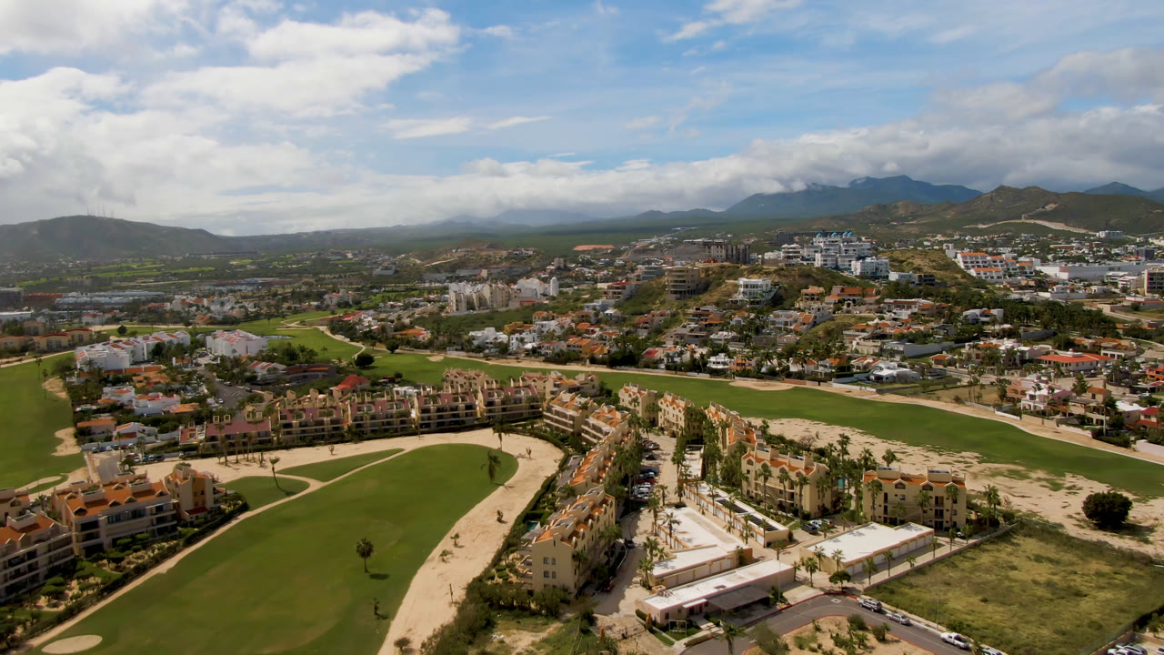 Aerial view over a golf course, buildings, roads, and mountains under clouds.