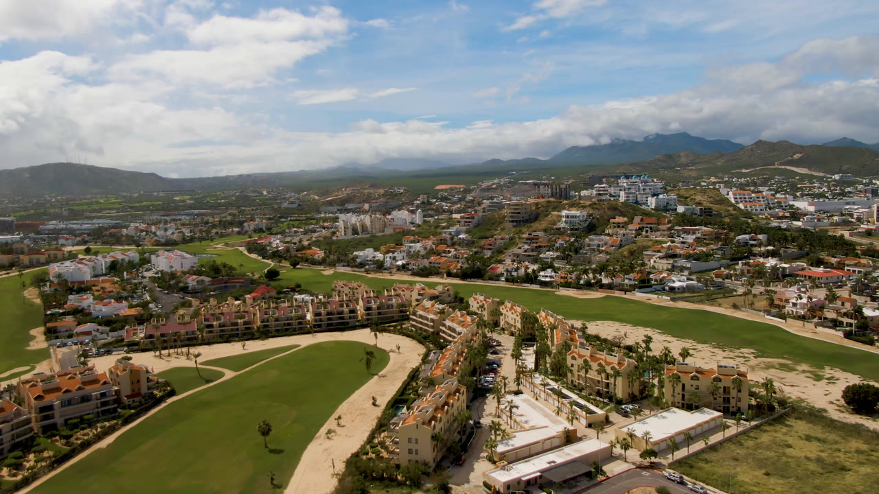 Aerial view in San Jose del Cabo with the fairway and pale sand areas across the lower frame, dense buildings in the middle distance, clouded mountains beyond, and no visible people.